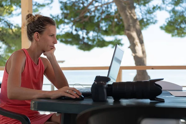 Young female photographer checking and editing photos on her laptop computer, sitting at a desk outside in summer resort.