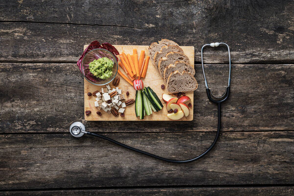 Top view of delicious and healthy vegan snack of sourdough bread, raw vegetables, tofu cubes and avocado spread served on wooden board. With medical stethoscope next to it.