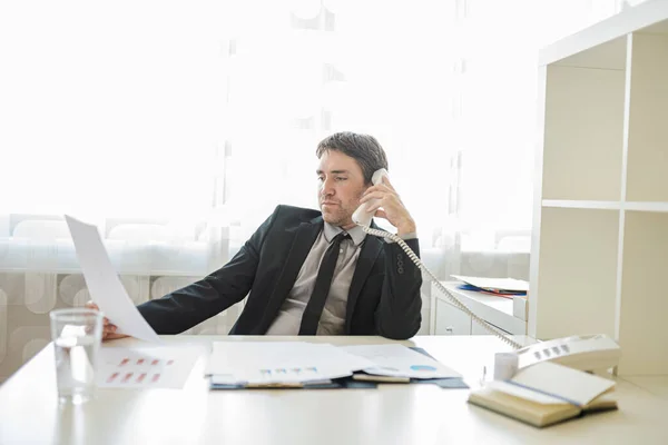 彼はレポートや書類を見て相談しながら 彼のオフィスの机に座って成功した若いビジネスマンは 電話で話しています — ストック写真