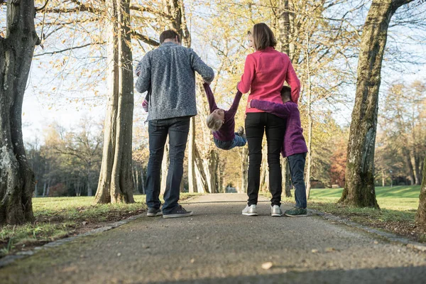 公園を歩いている間に手を握っている3人の子供を持つ若い家族の後ろからの眺め 真ん中でお母さんとお父さんが子供を持ち上げる — ストック写真