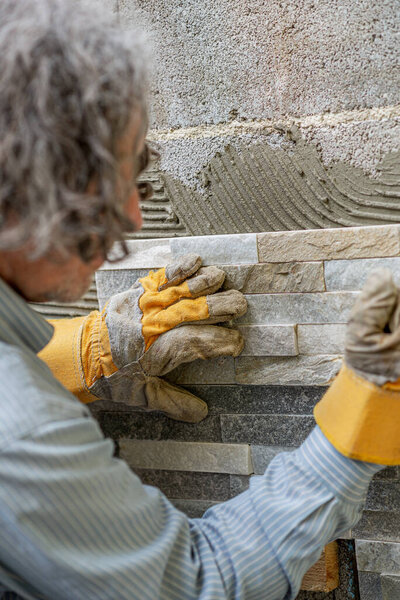 Senior man wearing protective workwear gloves as he presses ornamental tile in a glue on outside wall.