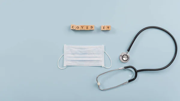 医療用保護マスクと聴診器の横にある木製のブロックに綴じ込まれたCovid 19サイン ライトブルーを背景に — ストック写真