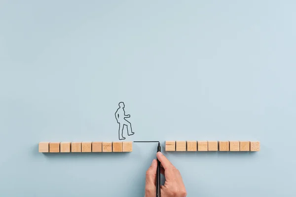 Male hand drawing a bridge between two rows of wooden blocks for a silhouetted man to walk across. Over blue background with plenty of copy space.