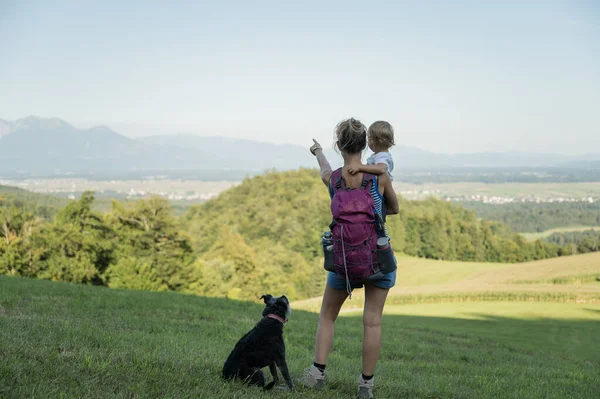 从一个背着远足背包的年轻母亲身后看去 她那蹒跚学步的女儿正站在美丽的草地上 指向美丽的风景 他们的黑狗坐在他们旁边 — 图库照片