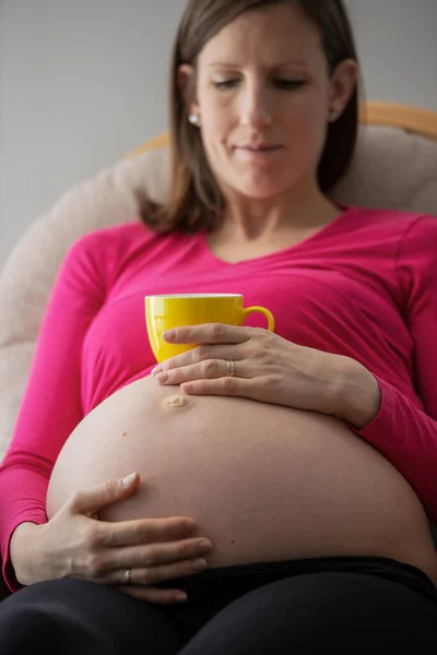 Pregnant young woman touching her barre swollen belly holding a cup of tea on top of it.