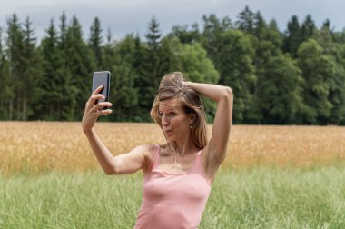 Genç bir kadın cep telefonuyla selfie çekerken komik bir yüz ifadesi takınıyor. Güzel yaz doğasında arka planda çayır ve tarla ile birlikte duruyor..