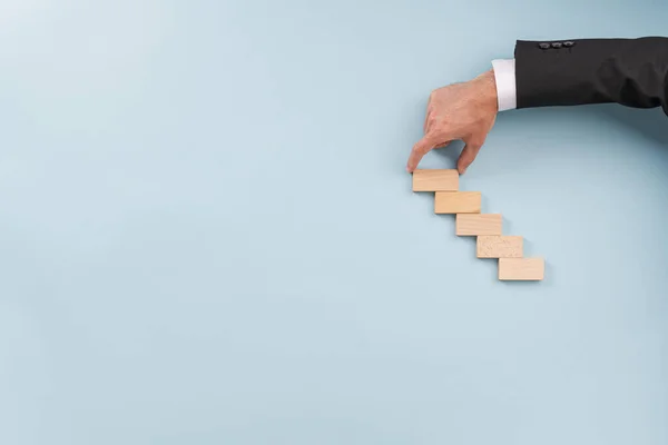 Hand of a businessman making stairway of wooden pegs over blue background with plenty of copy space.