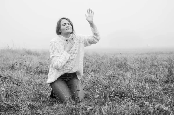 Girl closed her eyes on the knees, praying in a field during beautiful fog. Stock Photo