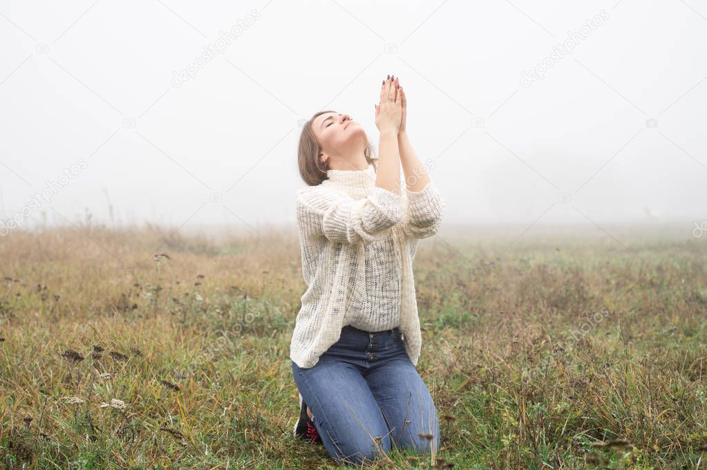 Girl closed her eyes on the knees, praying in a field during beautiful fog. 