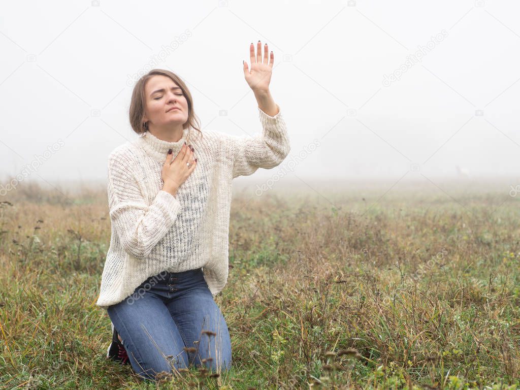 Girl closed her eyes on the knees, praying in a field during beautiful fog. 
