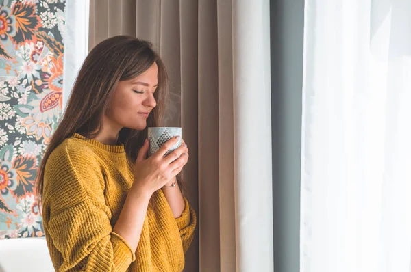 Femme avec une tasse de boisson chaude près de la fenêtre. En regardant fenêtre et boire du thé . — Photo