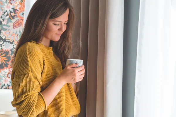 Mujer con una taza de bebida caliente junto a la ventana. Mirando a la ventana y beber té . — Foto de Stock