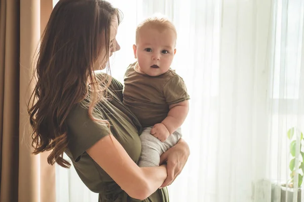 Retrato casero de un bebé con madre cerca de la ventana. Mamá sosteniendo y besando a su hijo . — Foto de Stock