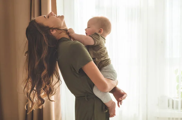 Házi portré egy kisfiúról anyával az ablak mellett. Anya öleli és csókolja a gyermekét. — Stock Fotó