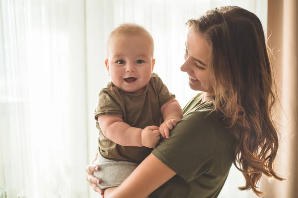 Potret rumah seorang bayi laki-laki dengan ibu dekat jendela. Ibu memegang dan mencium anaknya . — Stok Foto