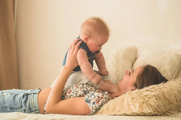 Szeretet és család. Házi portré egy kisfiúról anyával az ágyon. Anya játszik és megcsókolja a gyerekét. — Stock Fotó