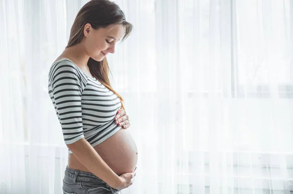 Feliz mujer embarazada con el vientre grande junto a la ventana. Conceptos de embarazo y familia — Foto de Stock