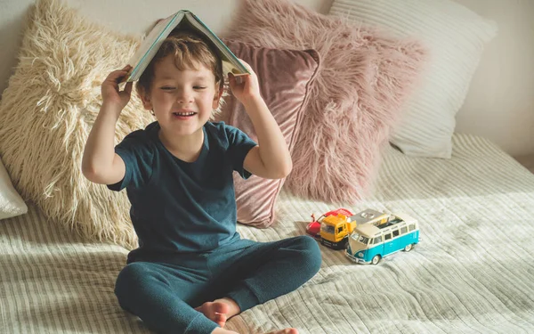 Un niño pequeño se sienta en una cama con sus juguetes en la sala de estar viendo imágenes en el libro de cuentos —  Fotos de Stock