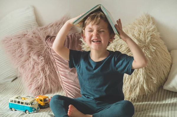 Un niño pequeño se sienta en una cama con sus juguetes en la sala de estar viendo imágenes en el libro de cuentos — Foto de Stock