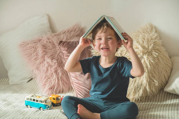 Un niño pequeño se sienta en una cama con sus juguetes en la sala de estar viendo imágenes en el libro de cuentos —  Fotos de Stock