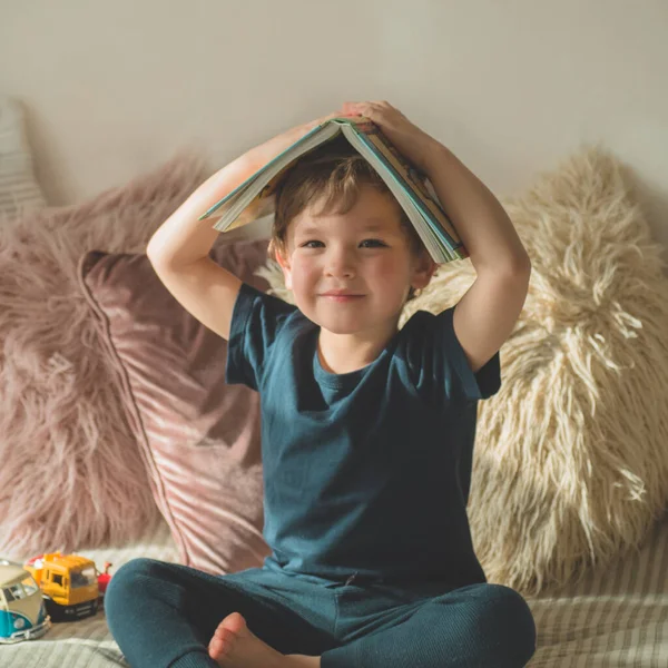 Un niño pequeño se sienta en una cama con sus juguetes en la sala de estar viendo imágenes en el libro de cuentos —  Fotos de Stock