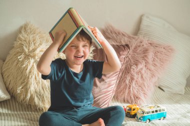 A little boy sits on a bed with your toys in living room watching pictures in story book