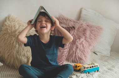 A little boy sits on a bed with your toys in living room watching pictures in story book