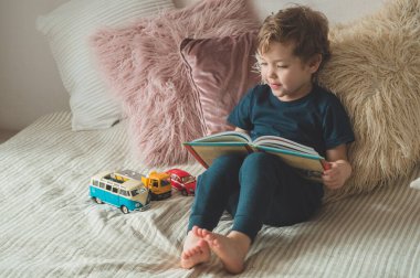 A little boy sits on a bed with your toys in living room watching pictures in story book