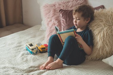 A little boy sits on a bed with your toys in living room watching pictures in story book
