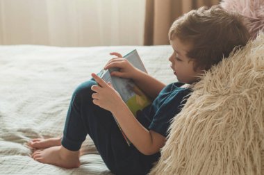 A little boy sits on a bed with your toys in living room watching pictures in story book
