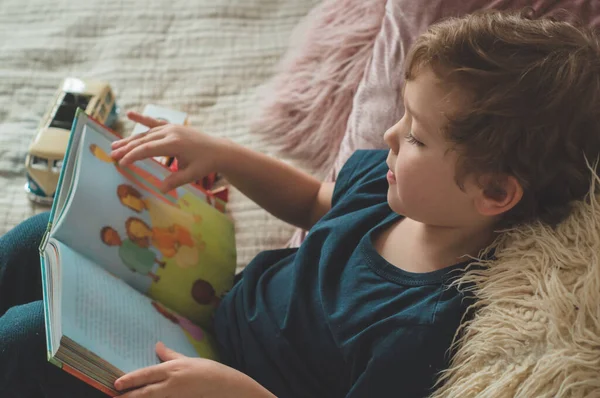 เด็กน้อยนั่งบนเตียงกับของเล่นของคุณในห้องนั่งเล่นดูรูปในหนังสือเรื่องราว — ภาพถ่ายสต็อก