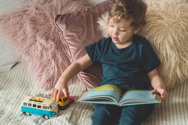 Um menino se senta em uma cama com seus brinquedos na sala de estar assistindo fotos no livro de histórias — Fotografia de Stock