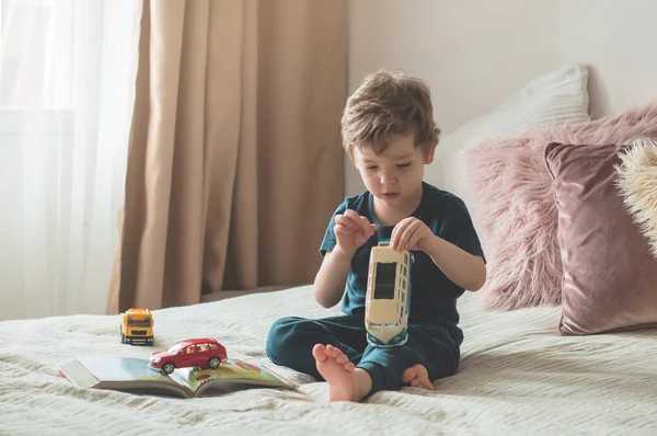 Un niño pequeño se sienta en una cama con sus juguetes en la sala de estar viendo imágenes en el libro de cuentos —  Fotos de Stock