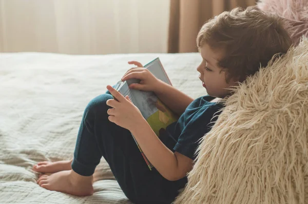 Un niño pequeño se sienta en una cama con sus juguetes en la sala de estar viendo imágenes en el libro de cuentos — Foto de Stock