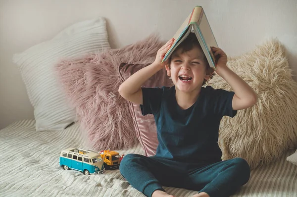 Un niño pequeño se sienta en una cama con sus juguetes en la sala de estar viendo imágenes en el libro de cuentos — Foto de Stock