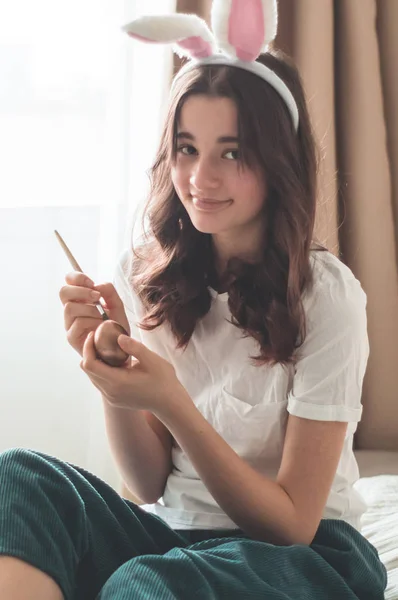 Chica adolescente con orejas de Pascua y pinta huevos de Pascua con un pincel en una cama en una sala de estar . — Foto de Stock