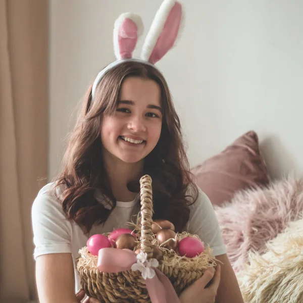 Tini lány húsvéti fülekkel és egy fonott húsvéti kosárral az ágyon a nappaliban — Stock Fotó