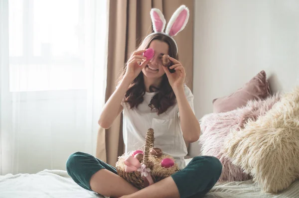 Adolescente avec des oreilles de Pâques et un panier de Pâques en osier ferme les yeux avec des œufs sur un lit dans un salon . — Photo