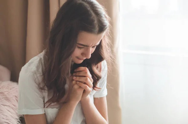 เด็กสาววัยรุ่นปิดตาของเธอ อธิษฐานในห้องนั่งเล่น มือพับในแนวคิดการสวดมนต์เพื่อความเชื่อ — ภาพถ่ายสต็อก