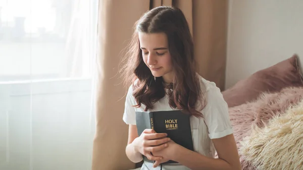 La adolescente cristiana tiene la Biblia en sus manos. Leyendo la Biblia en el salón. concepto de fe, espiritualidad y religión — Foto de Stock