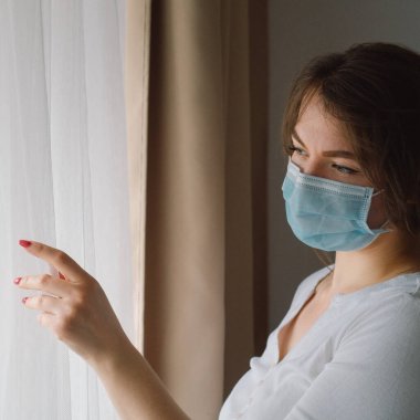 Coronavirus covid-19. Koruyucu maskeli bir kadın pencerenin yanında duruyor.