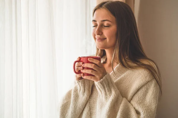 Mujer con una taza de bebida caliente junto a la ventana. Buenos días con el té. Otoño Invierno . — Foto de Stock