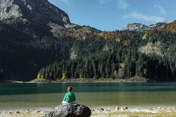 Büyük bir kaya üzerinde oturan çocuk ve sonbahar Kara Göl manzarasına sahiptir, — Stok fotoğraf