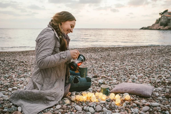 Jonge vrouw met grijze vacht met thee op het strand tijdens zonsondergang — Stockfoto