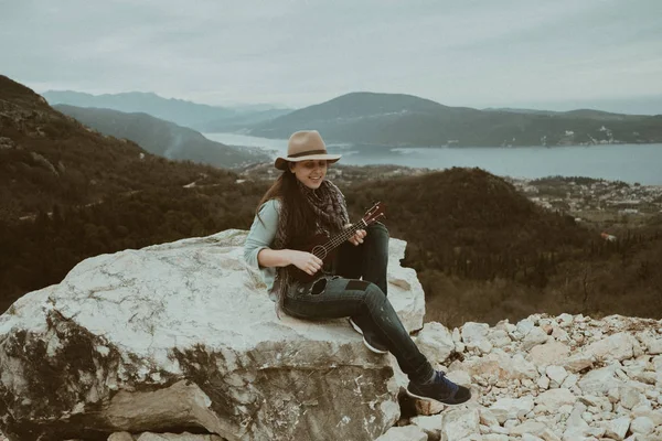 Gülümseyen şapkalı ukulele dağda oynayan bir genç kadın — Stok fotoğraf