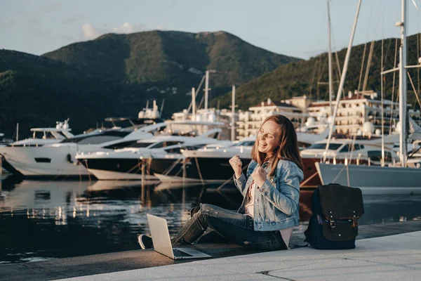 Vrolijke vrouw vrouw zittend op de dijk bij zonsondergang met laptop. — Stockfoto