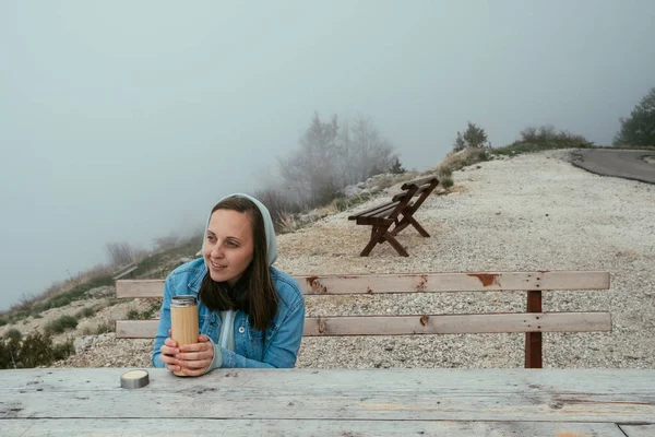 Jovem mulher sentada no banco de madeira e beber chá quente de um — Fotografia de Stock