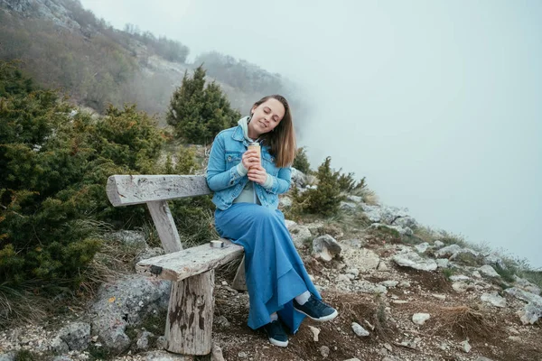 Mujer sonriente sentada en un banco de madera y bebiendo té caliente de —  Fotos de Stock