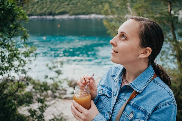 Jovem mulher bebe suco de laranja em um contexto de um mar landsca — Fotografia de Stock