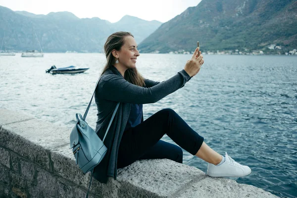 Charming woman in gray cardigan and blue backpack does selfie on — Stock Photo, Image